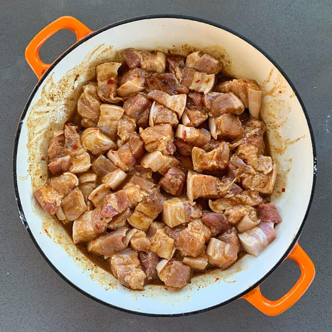 Sticky Chinese Pork Belly A round white pot with orange handles filled with cubed, marinated raw pork belly pieces sits on a gray surface.