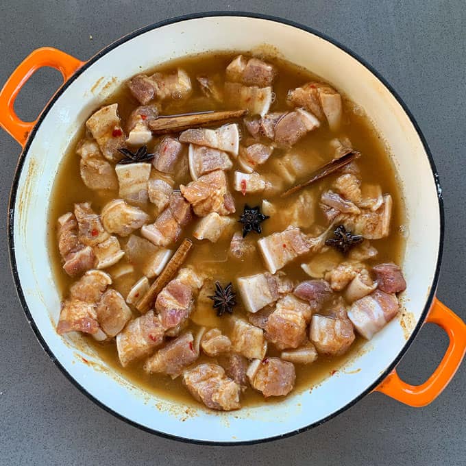 Sticky Chinese Pork Belly A pot with chunks of pork belly marinating in a brown liquid, surrounded by visible cinnamon sticks and star anise, sits on a gray countertop.