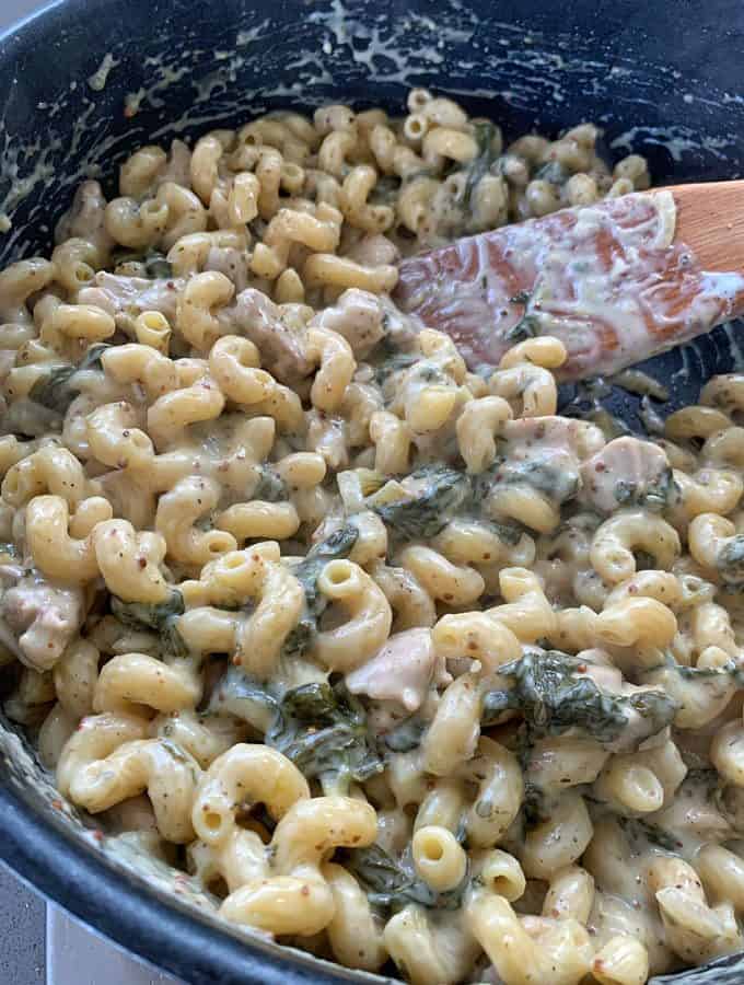 One pot chicken and pasta A close-up of one pot chicken pasta, with creamy sauce, pieces of chicken, and spinach mixed together in a pan, being stirred with a wooden spatula.