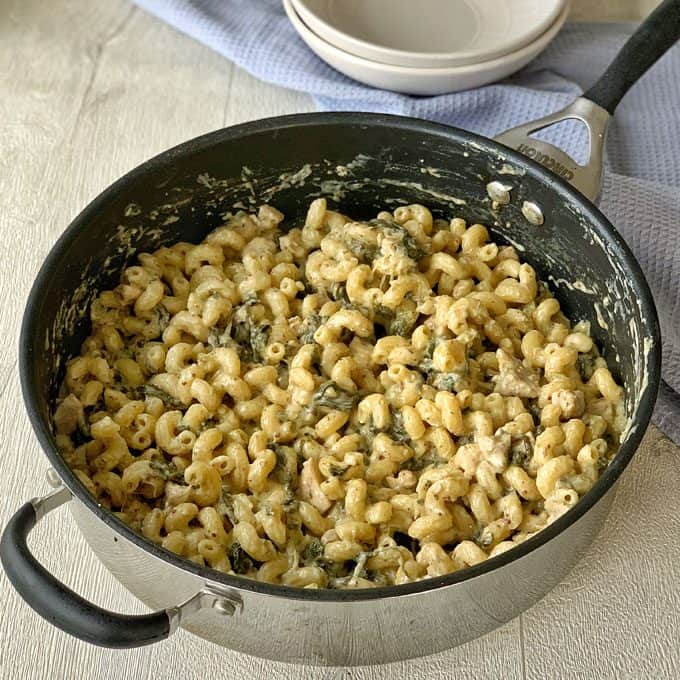 One pot chicken and pasta A pan filled with creamy one pot chicken pasta, featuring tender pieces of chicken and spinach, sits on a light wooden surface near stacked plates.