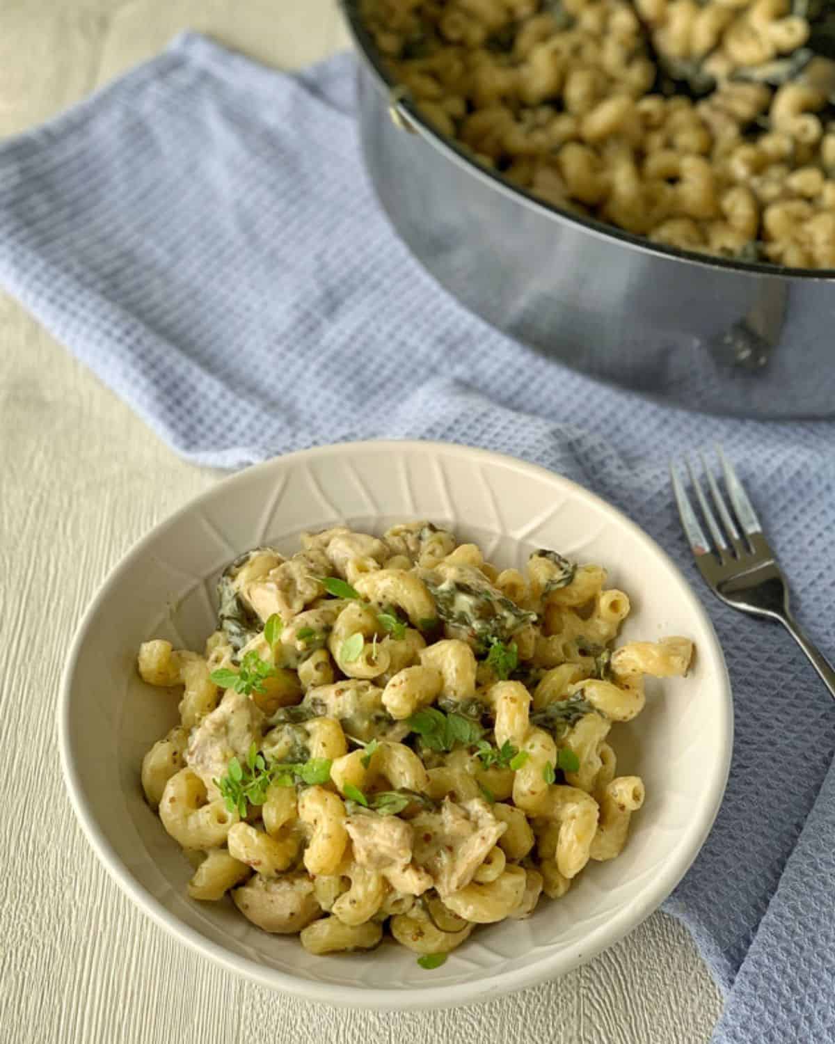 A bowl of creamy one pot chicken pasta with herbs sits on a table next to a fork and a pot of more pasta, all placed on a light blue textured cloth.