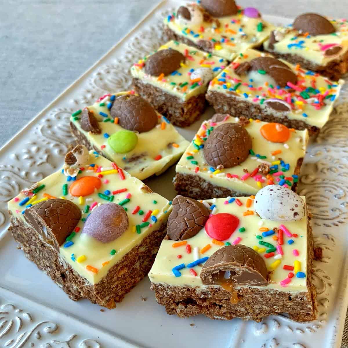 Easter Egg Slice A platter of chocolate-topped dessert bars with white chocolate, colorful sprinkles, Easter egg slices, and candy pieces.