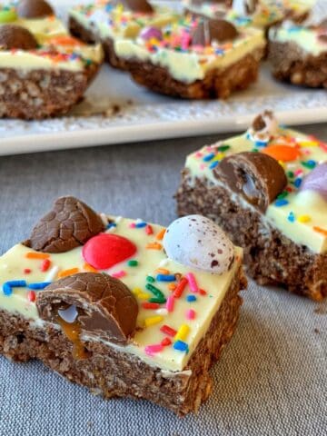 Close-up of colorful Easter egg slice squares topped with white icing, chocolate pieces, candy eggs, and rainbow sprinkles on a gray fabric surface, with more delectable squares on a white plate in the background.