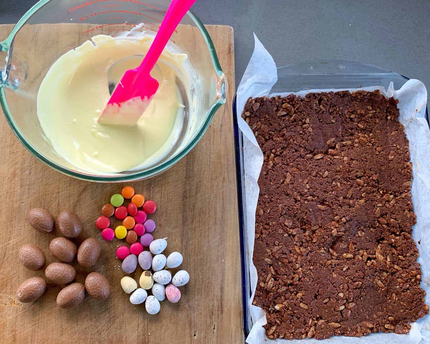 Easter Egg Slice A glass dish with chocolate mixture on the right, a glass bowl of melted white chocolate with a pink spatula on the left. Assorted chocolate eggs and candies, reminiscent of an Easter egg slice, are artfully arranged next to them.