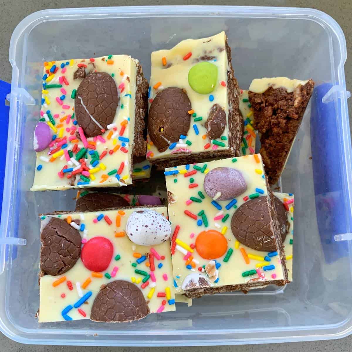 Easter Egg Slice Colorful Easter egg slices with white icing, topped with chocolate eggs, sprinkles, and candy, arranged in a clear plastic container.