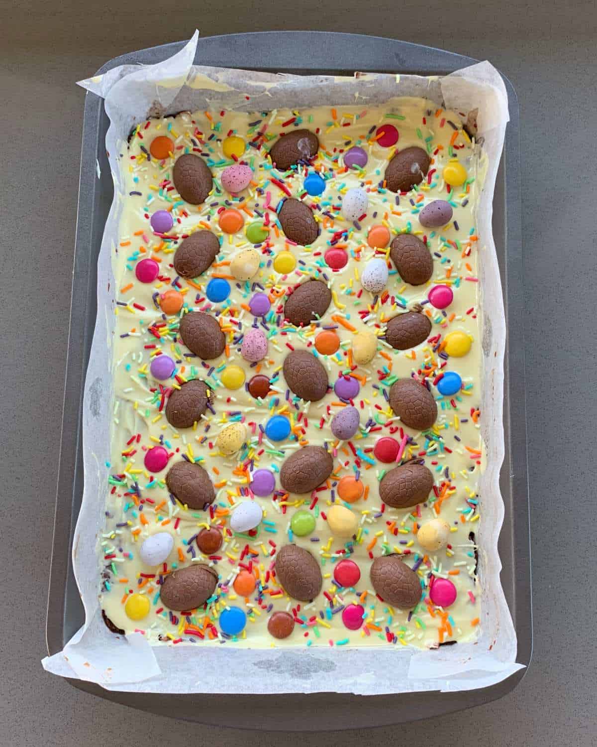 Easter Egg Slice A tray of colorful Easter egg slice treats featuring chocolate candies and sprinkles atop a layer of white coating, all arranged on a baking sheet lined with parchment paper.