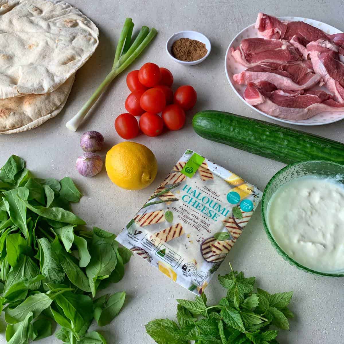 Lamb chops, cherry tomatoes, flatbread, spring onions, cucumber, lemon, garlic, cumin, yogurt, mint, arugula, and packaged halloumi cheese are arranged on a countertop—everything you need for delicious lamb and haloumi wraps.