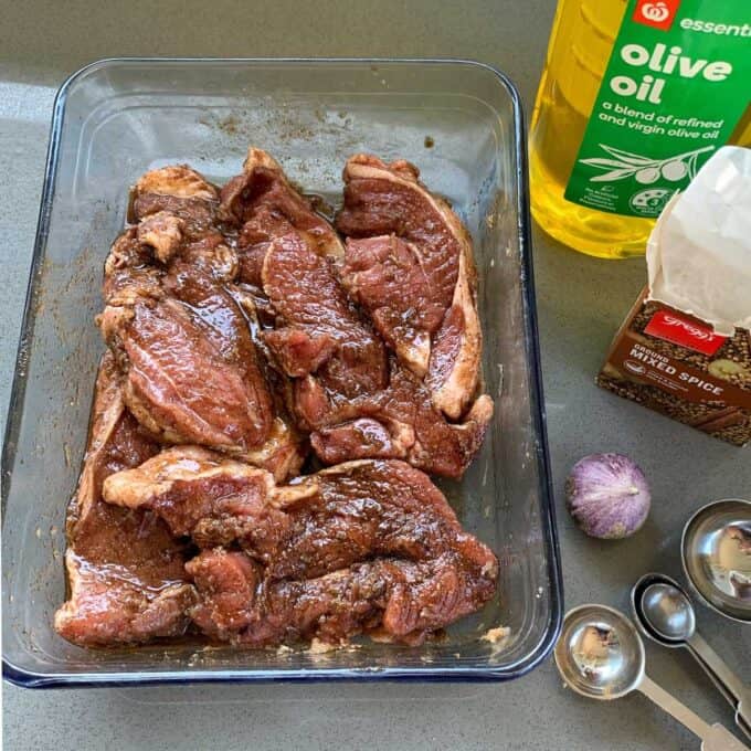Marinated meat pieces in a glass dish next to a bottle of olive oil, a packet of mixed spice, a shallot, and measuring spoons on a countertop—perfect for preparing delicious lamb and haloumi wraps.