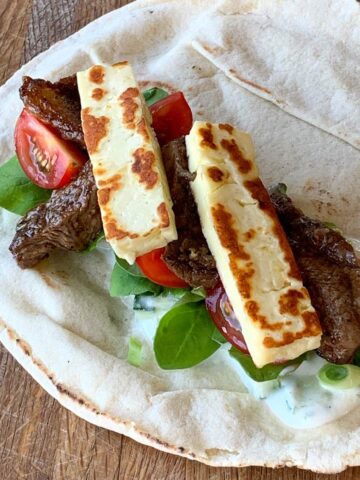 A flatbread topped with grilled halloumi cheese, beef strips, cherry tomatoes, spinach leaves, and a yogurt sauce—perfect for fans of lamb and halloumi wraps—placed on a wooden surface.