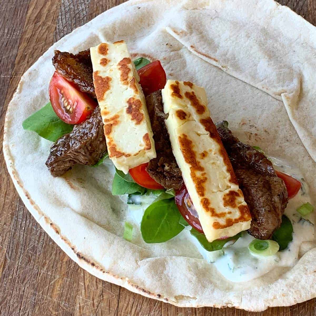A flatbread topped with grilled halloumi cheese, beef strips, cherry tomatoes, spinach leaves, and a yogurt sauce—perfect for fans of lamb and halloumi wraps—placed on a wooden surface.