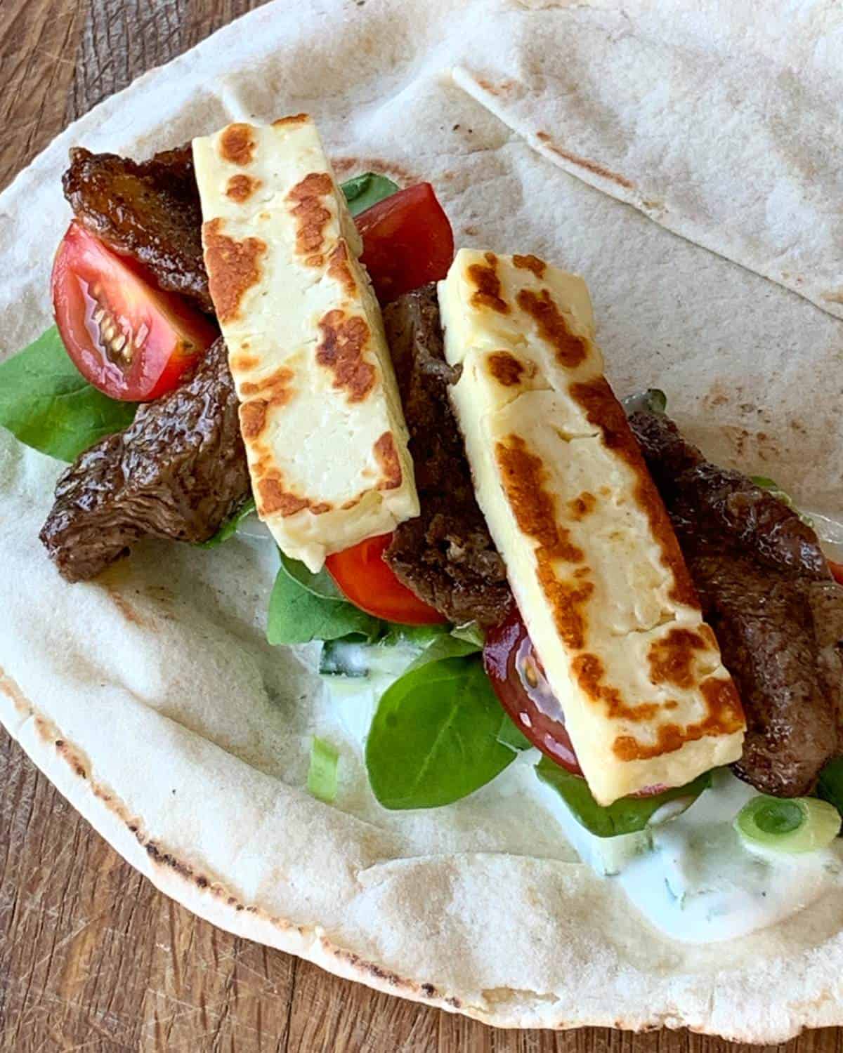 Flatbread topped with grilled halloumi, strips of cooked beef, fresh spinach, cherry tomatoes, and creamy white sauce—a delicious twist inspired by Lamb and Haloumi Wraps. Served on a wooden surface.