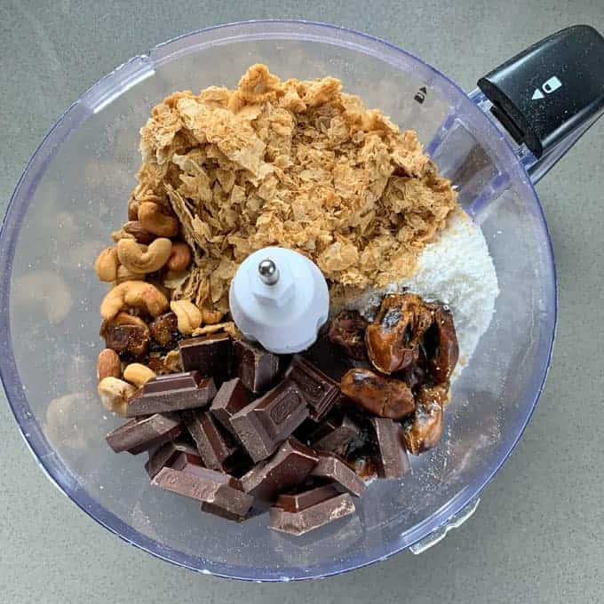 A food processor bowl filled with chocolate chunks, cashews, shredded coconut, dates, and cereal flakes—perfect for making Weetbix bliss balls, ready to be blended into a delicious treat.
