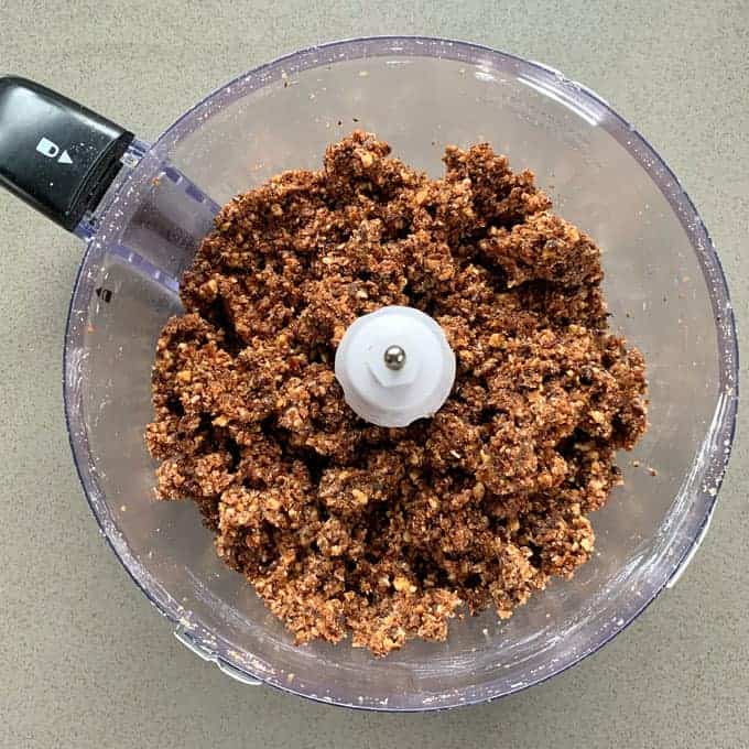 A food processor bowl containing a coarse, mixed brown and beige crumbly mixture for Weetbix bliss balls sits on a gray countertop.