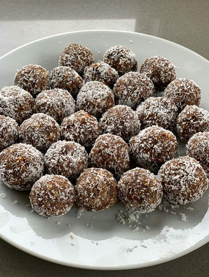 Chocolate, Date and Weetbix Bliss Balls A white plate filled with Chocolate Date Weetbix Bliss Balls coated in shredded coconut, arranged in rows on a light-colored surface.