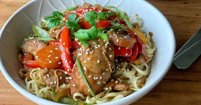 Slow Cooker Honey and Soy Chicken A white bowl filled with Slow Cooker Honey Soy Chicken, noodles, sliced carrots, red bell peppers, green onions, cilantro, and a sprinkle of sesame seeds on a wooden surface.