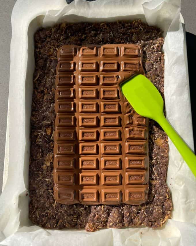 A baking tray lined with parchment paper holds a Chocolate Weetbix Slice oat and chocolate mixture, topped with a chocolate bar being spread out smoothly using a green spatula.
