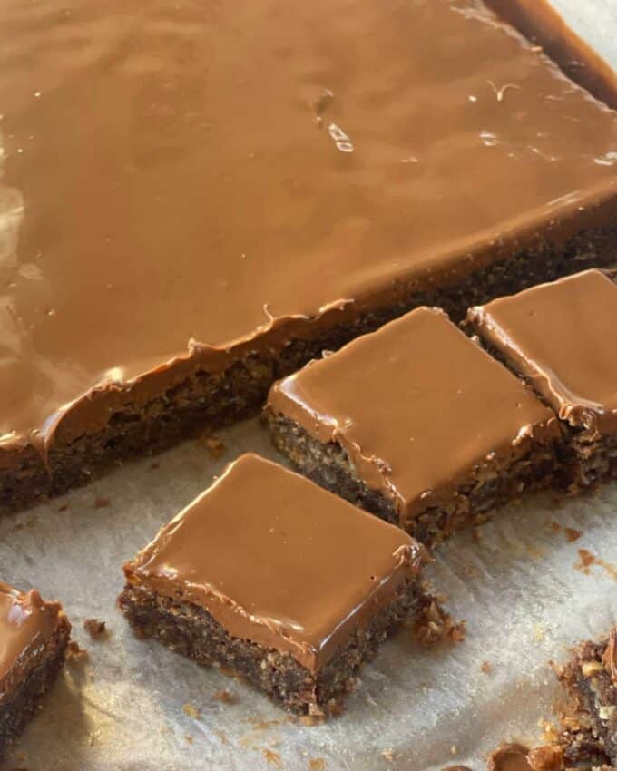A tray of Chocolate Weetbix Slice sheet cake with a glossy chocolate frosting, partially cut into square pieces, displayed on parchment paper.