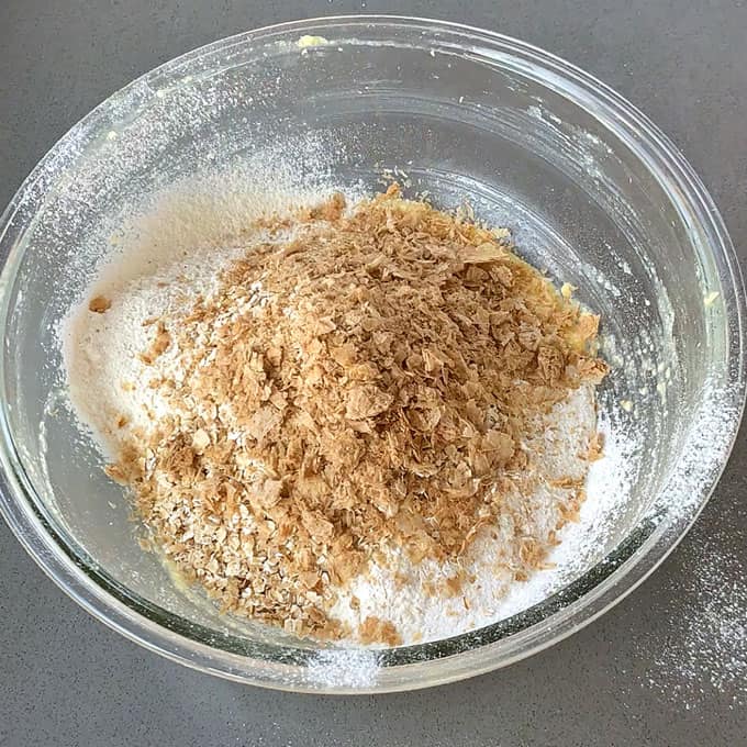 Flour, and crushed weetbix in a glass bowl on a grey bench.