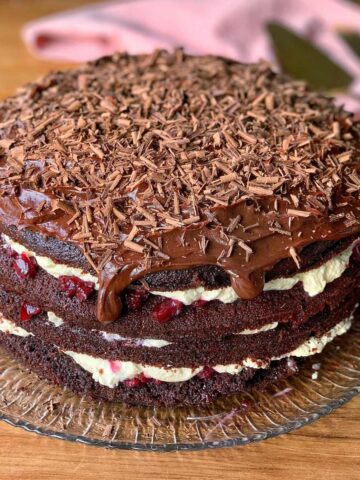 A luscious Black Forest Gateau, featuring layers of chocolate cake with cream and cherries nestled between, is elegantly topped with chocolate shavings. It sits invitingly on a glass plate, set against a backdrop of soft pink cloth.