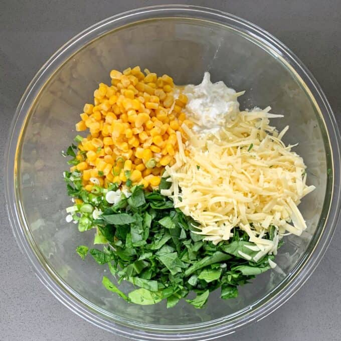 A glass bowl containing ingredients for corn fritters—corn, shredded cheese, chopped spinach, sliced green onions, and a dollop of mayonnaise or cream—sits on a gray surface.