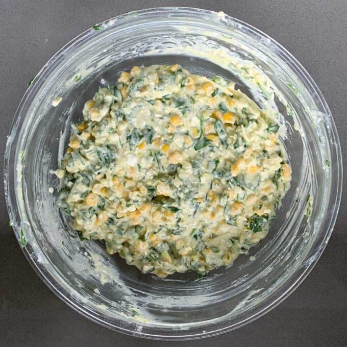 A glass bowl filled with a mixed batter for corn fritters, featuring corn, chopped greens, and a creamy, pale yellow mixture, placed on a grey surface.