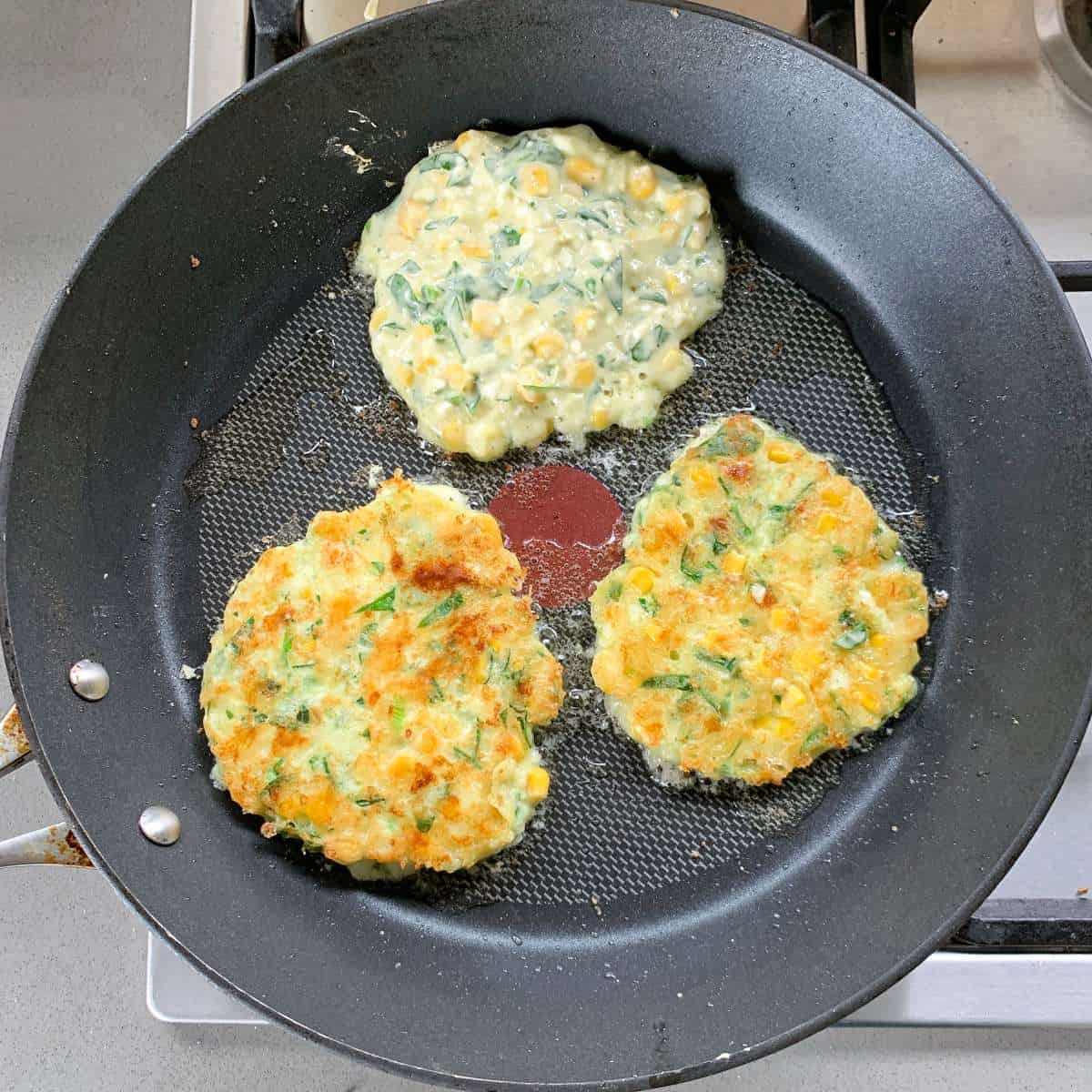 Three corn fritters with herbs are cooking in a non-stick frying pan; two corn fritters are golden brown, while the third remains raw and pale.