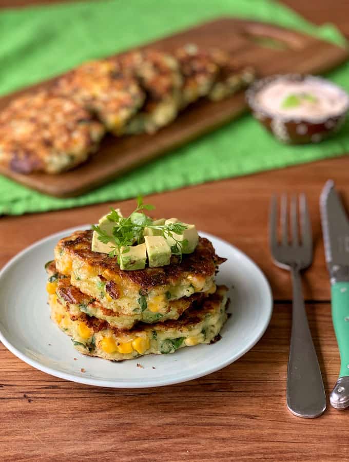 Three corn fritters topped with diced avocado and herbs are served on a plate, with a fork and knife beside it; more fritters and a dipping sauce appear in the background.