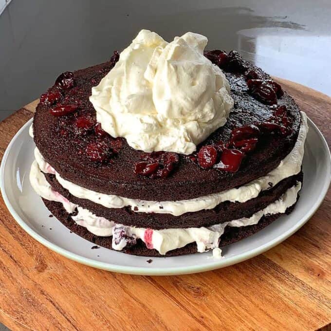 This Black Forest Gateau is a three-layer chocolate cake adorned with cherries and whipped cream between layers and on top, all beautifully presented on a white plate resting on a wooden surface.