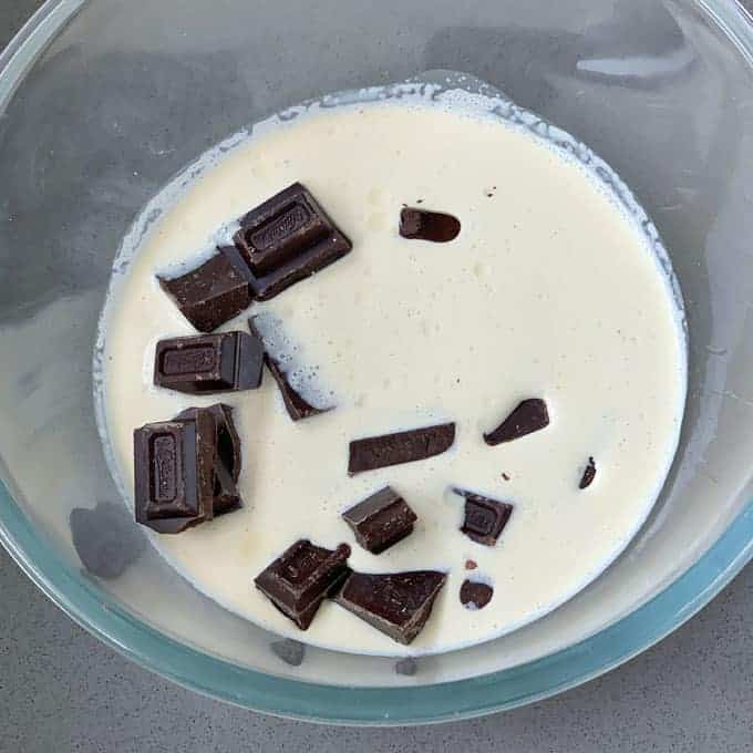 The glass bowl, reminiscent of a deconstructed Black Forest Gateau, holds silky cream and rich pieces of dark chocolate on a gray surface.