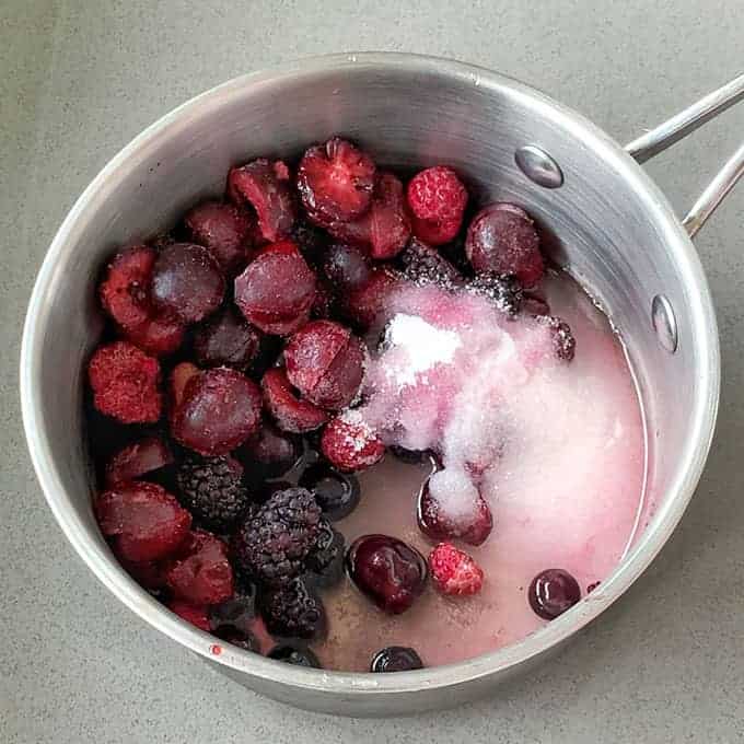 A saucepan with mixed berries, sugar, and liquid reminiscent of Black Forest Gateau sits enticingly on a gray surface.