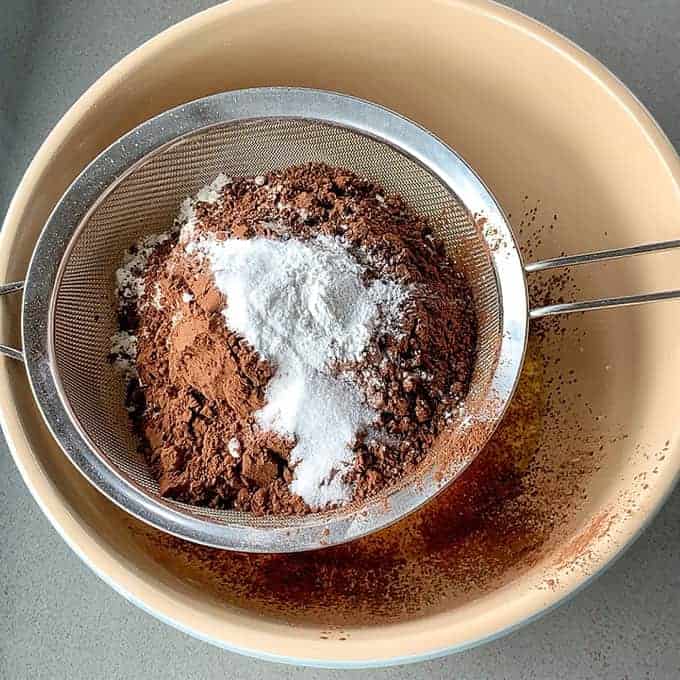 Brown and white powders, reminiscent of the rich layers of a Black Forest Gateau, are being sifted through a metal strainer over a beige bowl, likely cocoa powder and sugar.