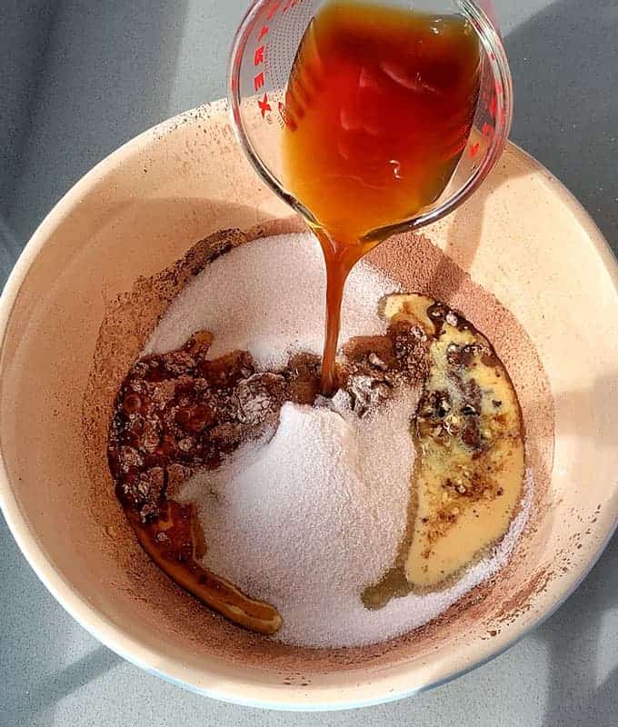 Pouring liquid into a mixing bowl with sugar, cocoa powder, flour, and other ingredients for a decadent Black Forest Gateau on a gray surface.