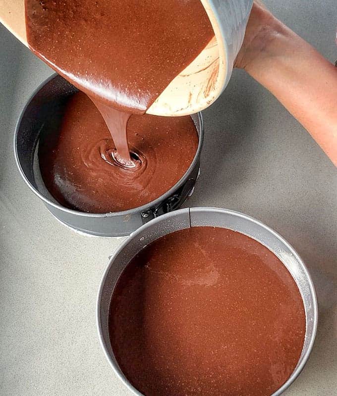 Pouring rich Black Forest Gateau batter into two round baking pans on a gray countertop.