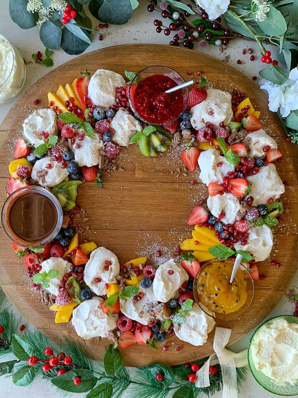 A birds eye view of a mini pavlova wreath with fruit dipping sauces on a circular wooden platter.