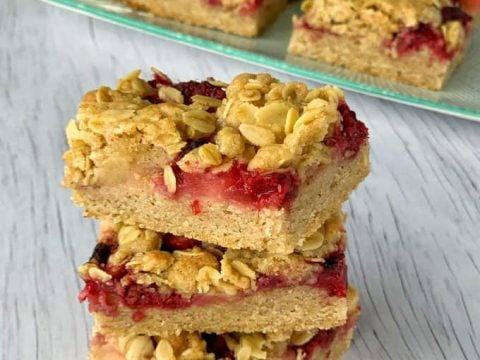 Three stacked apple and raspberry crumble slice bars sit in the foreground, with more bars on a plate in the background, all on a light wooden surface.