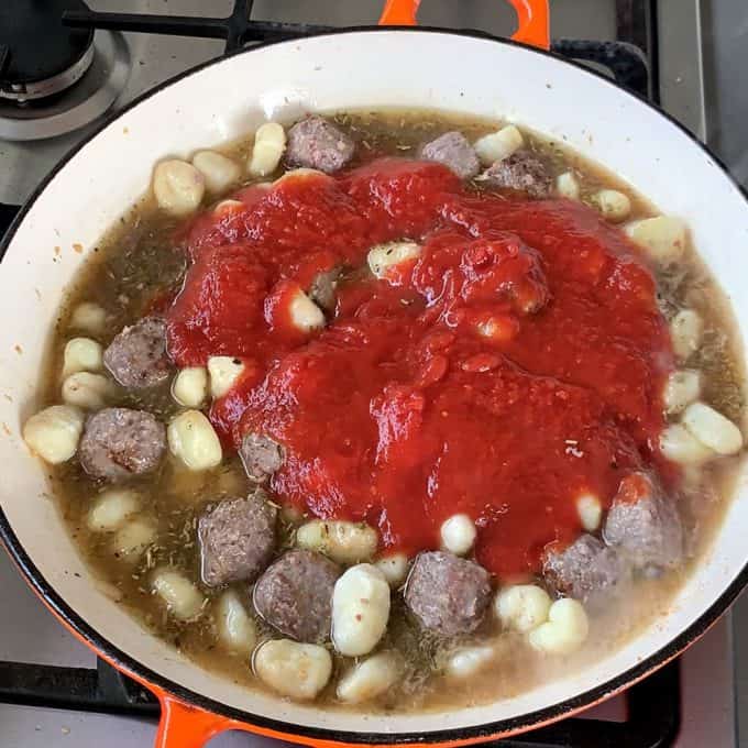A pan on a stove contains one pot meatballs and gnocchi simmering in broth and marinara sauce, partially cooked and filling the kitchen with a savory aroma.