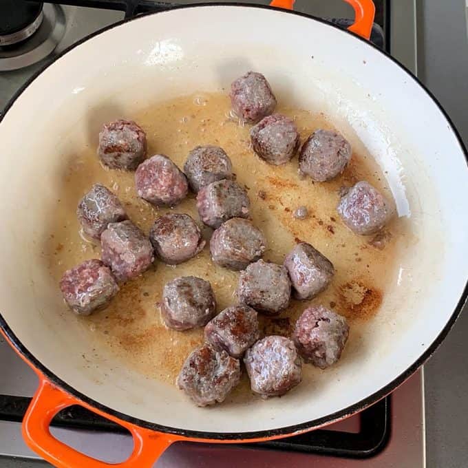 A white and orange pan on a stove contains several partially browned one pot meatballs and gnocchi cooking in oil.