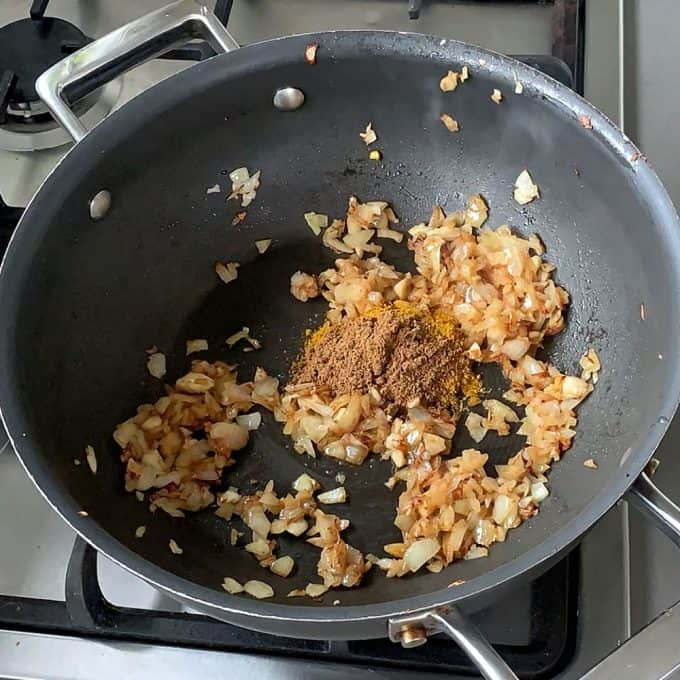 Chopped onions are being sautéed in a black pan on a stove, with a heap of ground spices added on top for a flavorful pumpkin and chickpea curry.