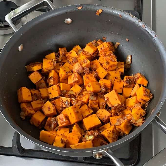 Cubed butternut squash with spices and onions cooking in a black pot on a stove, perfect for adding to a hearty pumpkin and chickpea curry.