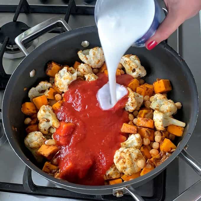 A hand pours coconut milk into a pan filled with cauliflower, sweet potatoes, chickpeas, and tomato sauce on a stovetop for a hearty pumpkin and chickpea curry.