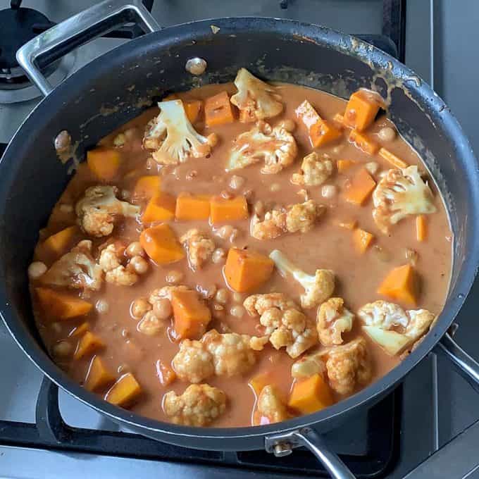 A black saucepan on a stove contains a pumpkin and chickpea curry with chunks of cauliflower, sweet potato, and beans in a creamy orange sauce.