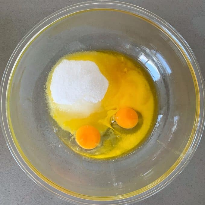 A clear glass bowl containing two raw eggs and a mound of white granulated sugar on a gray countertop, ready to be mixed for delicious banana muffins.