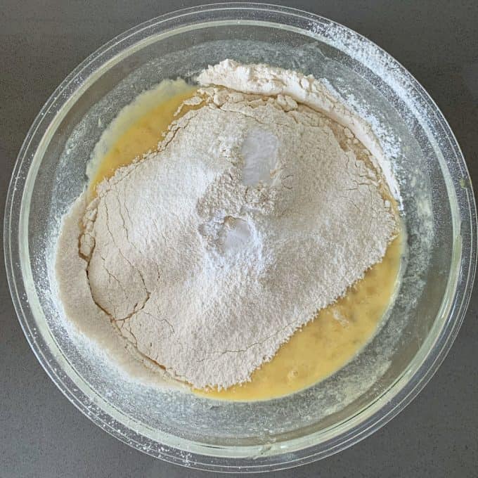 A glass bowl containing flour, baking powder, and a wet mixture for banana muffins batter sits on a gray countertop.
