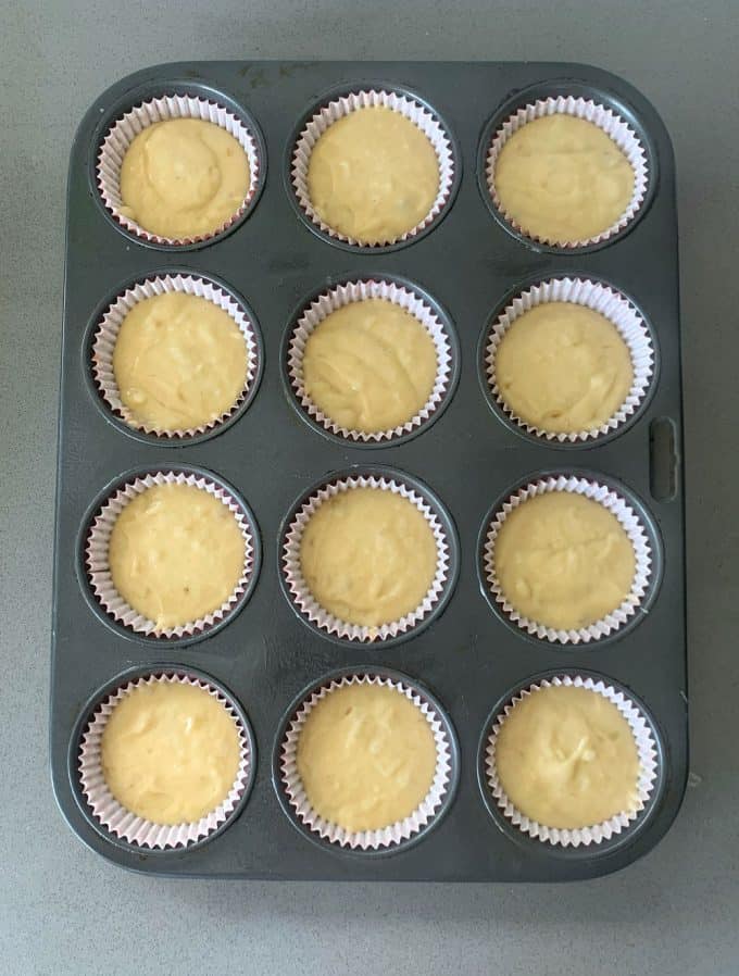A muffin tin with twelve paper liners, each filled with unbaked banana muffin batter, sits on a gray countertop.