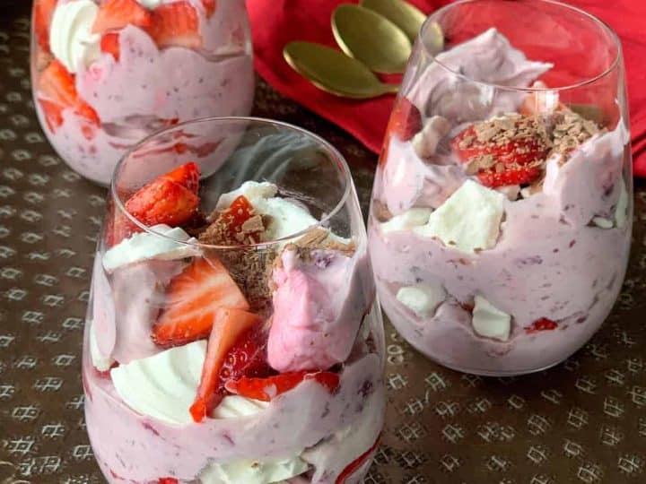 Three glasses filled with Ambrosia Eton Mess strawberry mousse dessert, topped with fresh strawberries and whipped cream, are arranged on a metal tray with a red napkin and two gold spoons.
