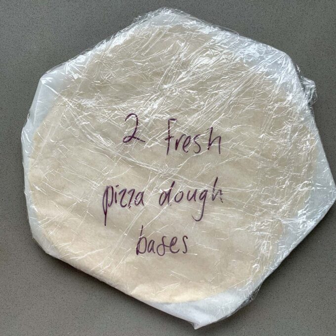 two pizza bases wrapped in cling film on a  grey bench.