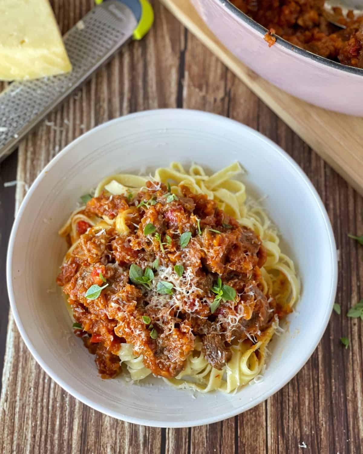 Oven Baked Beef Ragu A white bowl of tagliatelle pasta topped with oven baked beef ragu and tomato sauce, garnished with grated cheese and fresh herbs, sits on a wooden surface near a cheese grater.