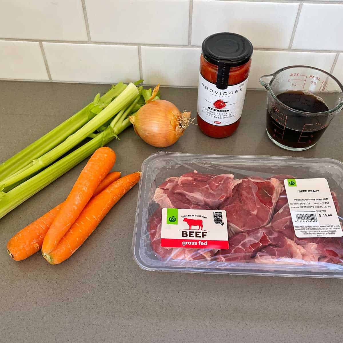 A package of grass-fed beef, three carrots, celery stalks, onion, tomato sauce, and beef gravy are ready on the kitchen counter for a delicious oven baked beef ragu.