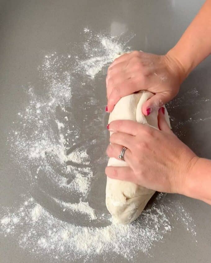 Two hands rolling pizza dough out on a floured grey bench top.