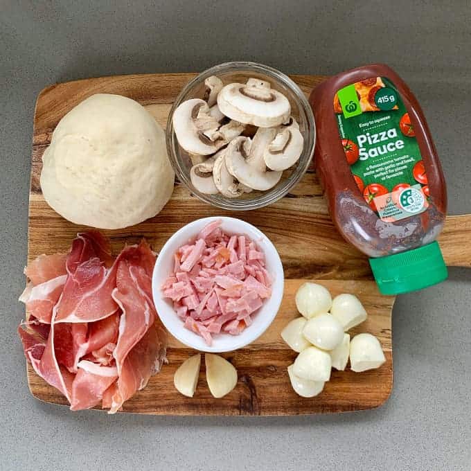 The ingredients to make a mushroom and prosciutto pizza on a wooden chopping board.